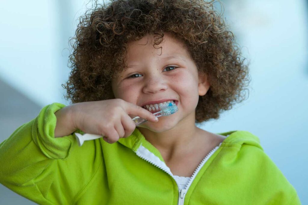 small child with curly hair looking smiling and brushing teeth