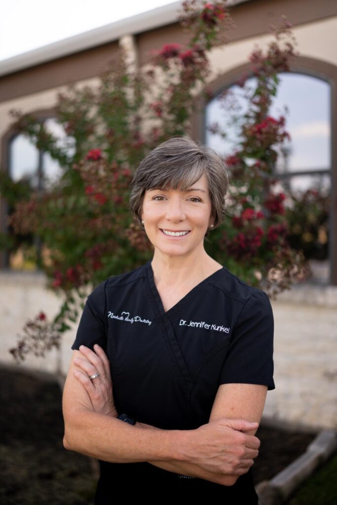 Dr. Jennifer Kunkel in black scrubs stands outdoors in front of a building and flowering tree, smiling with her arms crossed.