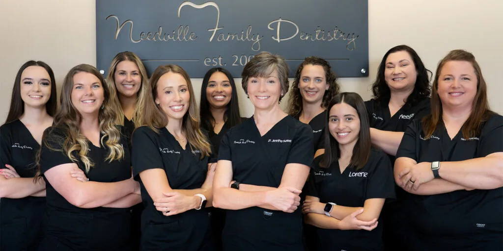 Group photo of ten smiling staff members from Needville Family Dentistry. All are dressed in black embroidered uniforms, posed with arms crossed or hands clasped, conveying a professional and welcoming team atmosphere inside the dental clinic.