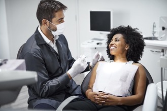 Dental professional wearing mask and gloves interacting with patient in chair with protective bib, surrounded by dental equipment during consultation.