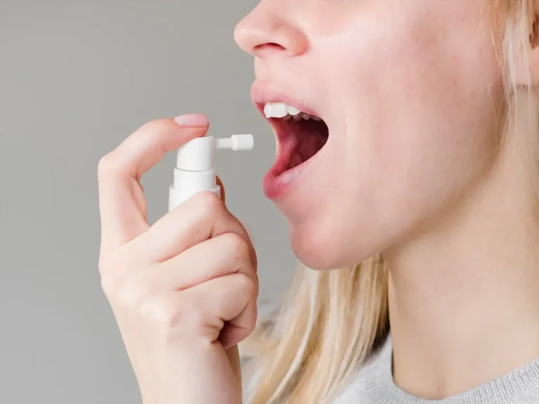 Person holding small spray bottle near open mouth, applying oral spray against plain background.