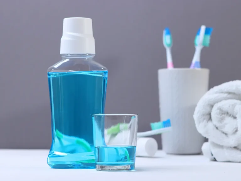 Bathroom setup with blue mouthwash bottle, glass of blue liquid, two toothbrushes in white holder, and rolled white towel on gray background.