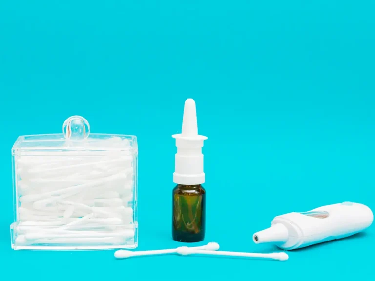 Transparent container of cotton swabs, brown nasal spray bottle, white electronic thermometer, and two loose swabs arranged against bright turquoise background.