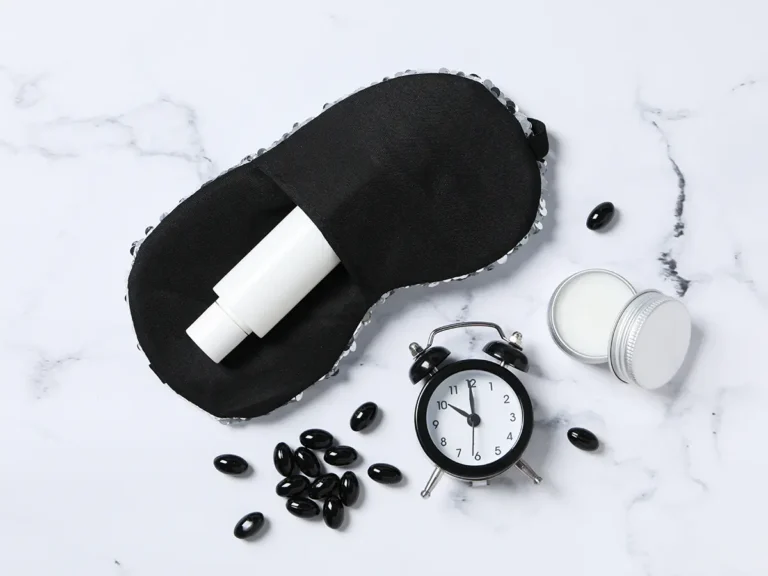 Flat lay on white marble surface with black sleep mask, cylindrical container, small black alarm clock, white jar, and scattered black capsules.